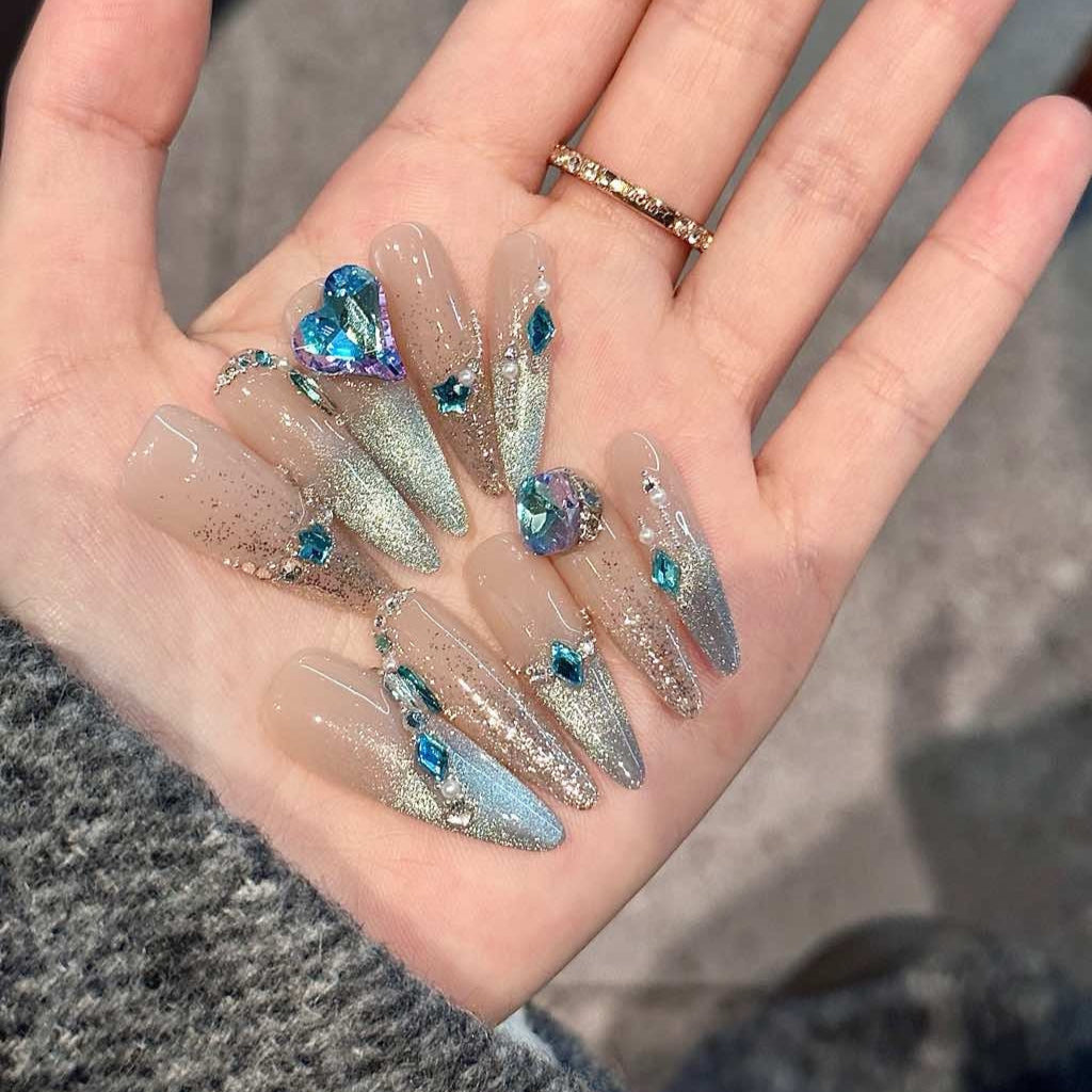 Embrace cosmic elegance with Celestial Frost Press-On Nails. These extra-long stiletto nails feature a stunning nude-to-silver gradient, accented with shimmering metallic details and radiant blue gemstones. The dazzling heart-shaped crystal centerpiece adds a touch of ethereal beauty, while delicate silver embellishments create a celestial, icy-glam aesthetic.