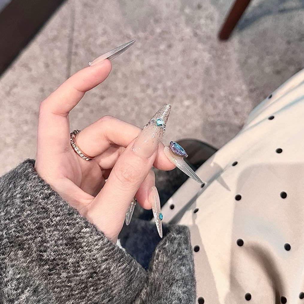 Embrace cosmic elegance with Celestial Frost Press-On Nails. These extra-long stiletto nails feature a stunning nude-to-silver gradient, accented with shimmering metallic details and radiant blue gemstones. The dazzling heart-shaped crystal centerpiece adds a touch of ethereal beauty, while delicate silver embellishments create a celestial, icy-glam aesthetic.