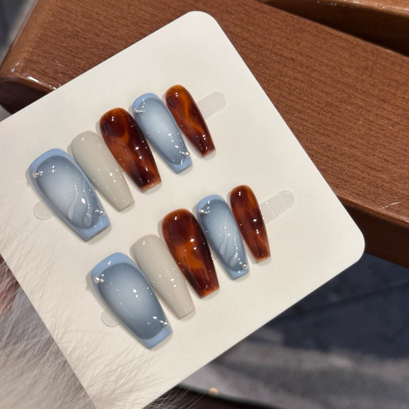 Embrace the contrast of cool elegance and warm intensity with Frost & Ember Press-On Nails. This striking set features a dual-tone design, blending icy blue cat-eye shimmer, creamy ivory, and deep amber brown for a bold yet sophisticated look. The sleek coffin shape adds a modern, elongated finish, while subtle rhinestone accents bring a touch of refined luxury.