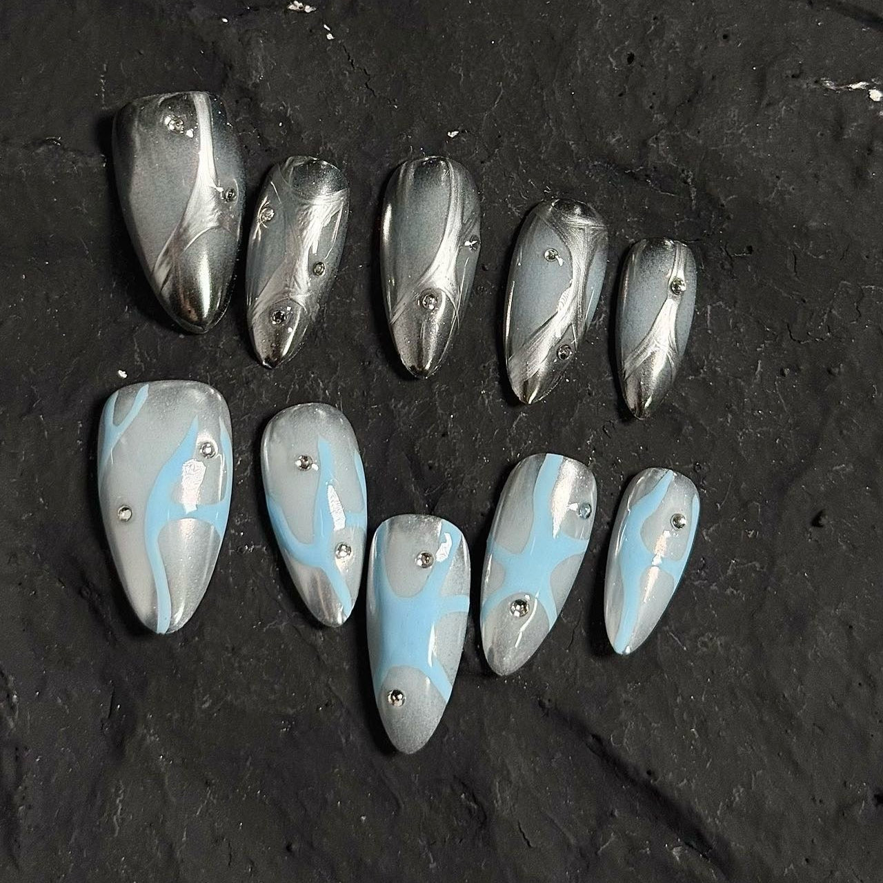 Frosted Waves Press-On Nails capture the serene elegance of icy tides with a metallic silver and cool blue color palette. Featuring fluid, wave-like patterns and delicate silver studs, these nails evoke a sense of calm yet futuristic sophistication.