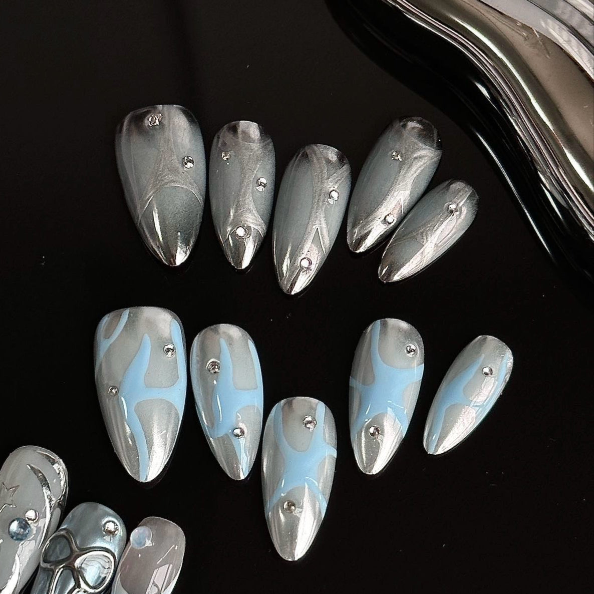 Frosted Waves Press-On Nails capture the serene elegance of icy tides with a metallic silver and cool blue color palette. Featuring fluid, wave-like patterns and delicate silver studs, these nails evoke a sense of calm yet futuristic sophistication.