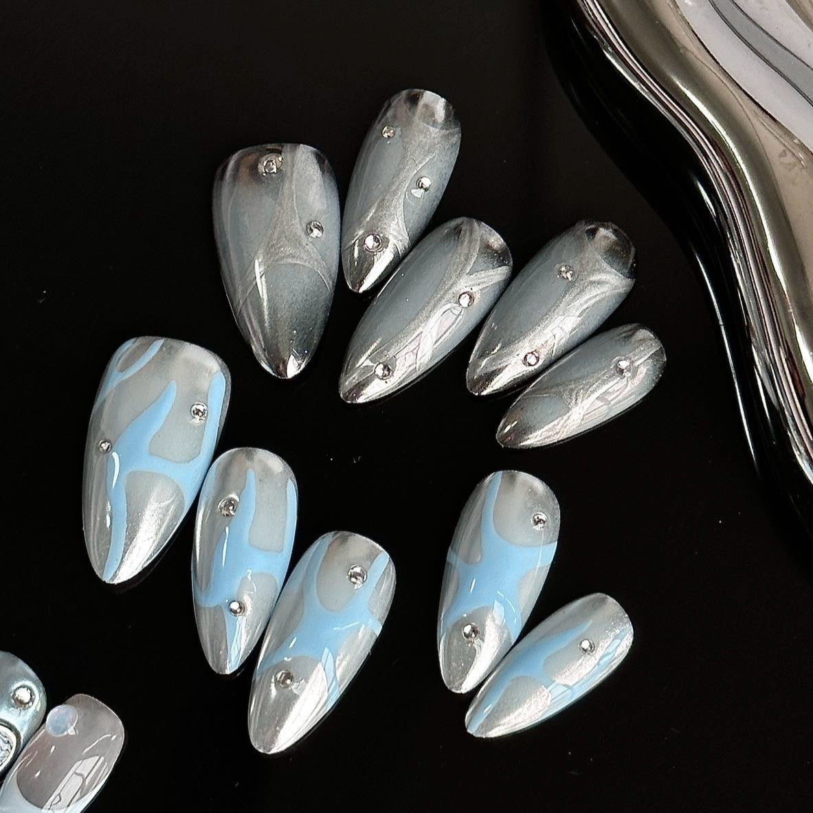 Frosted Waves Press-On Nails capture the serene elegance of icy tides with a metallic silver and cool blue color palette. Featuring fluid, wave-like patterns and delicate silver studs, these nails evoke a sense of calm yet futuristic sophistication.