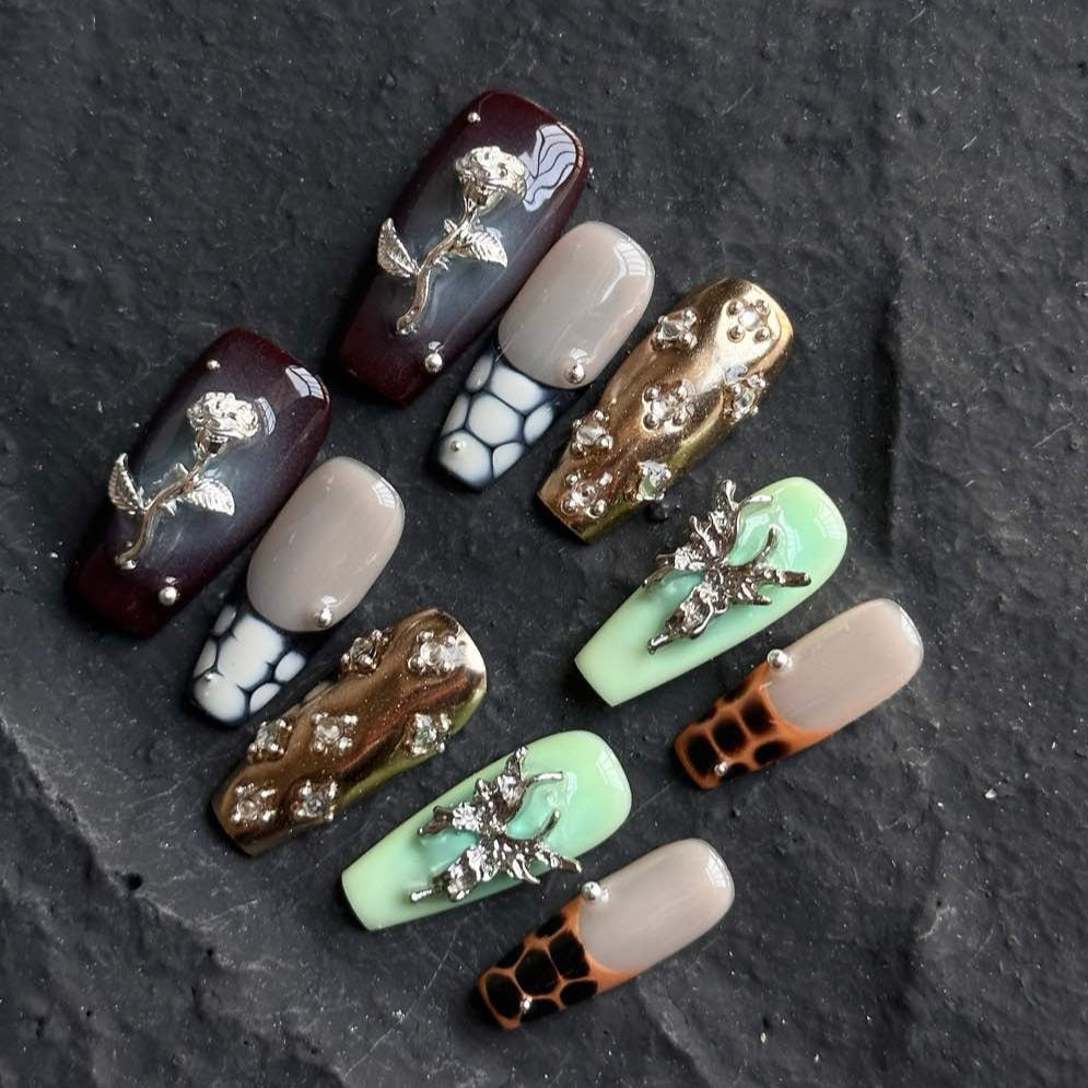 Channel bold luxury with Gilded Serpent Press-On Nails, an exotic fusion of rich jewel tones, metallic textures, and intricate embellishments. This opulent set features deep burgundy, jade green, and soft taupe hues, accentuated with snakeskin-inspired patterns, reflective gold chrome, and striking 3D silver charms. The blend of textures—smooth glossy finishes, sculpted reptilian details, and shimmering crystal accents—creates an edgy yet elegant masterpiece for your fingertips.