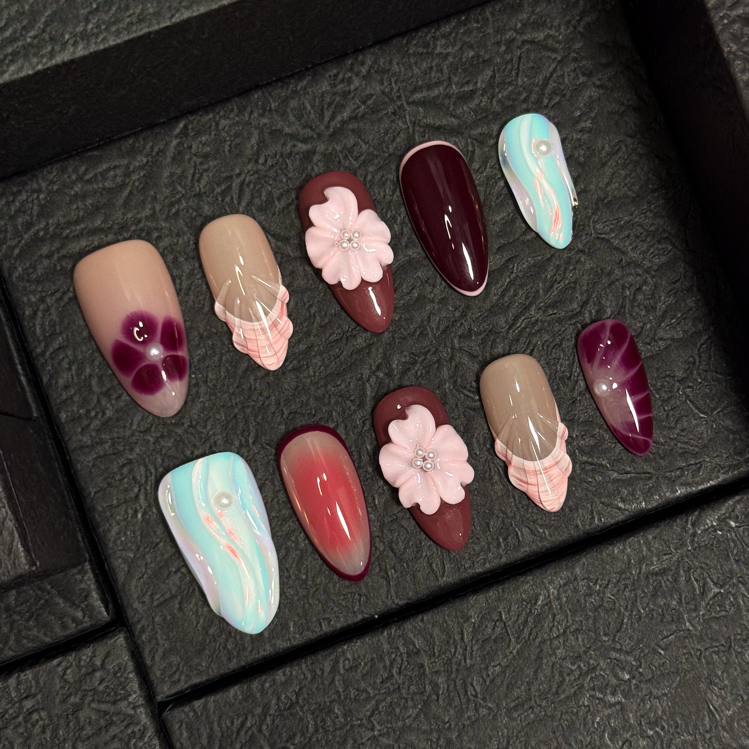 Rosy, Plum & Aqua Palette: A moody yet sweet mix of mauve, burgundy, shell beige, and ethereal pastel teal.
• 3D Blossoms & Shells: Hand-sculpted flowers and seashell ridges bring sculptural beauty and texture.
• Iridescent Swirls: Pastel wave patterns with a glowing pearl finish mimic soft ocean tides.
• Elegant Accents: Pearl beads and gradient effects add depth and femininity to each nail.
• Almond Shape: A gentle, elongated silhouette that enhances both softness and sophistication.