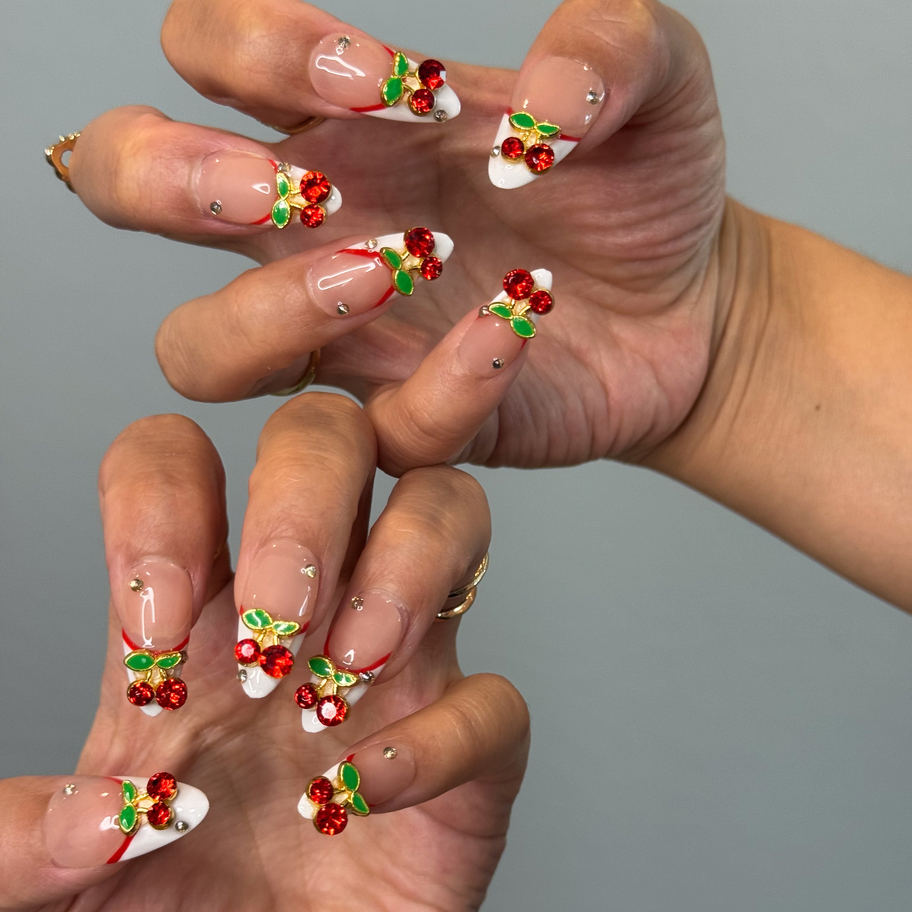 Red French Accent: Classic meets modern with a deep red curve layered over crisp white.
• 3D Cherry Charms: Crystal-studded cherries and metallic green leaves bring a touch of fruity glam.
• Nude Gloss Base: A soft, neutral backdrop keeps the design fresh and wearable.
• Gold Stud Details: Subtle metallic accents add a hint of luxe sparkle.