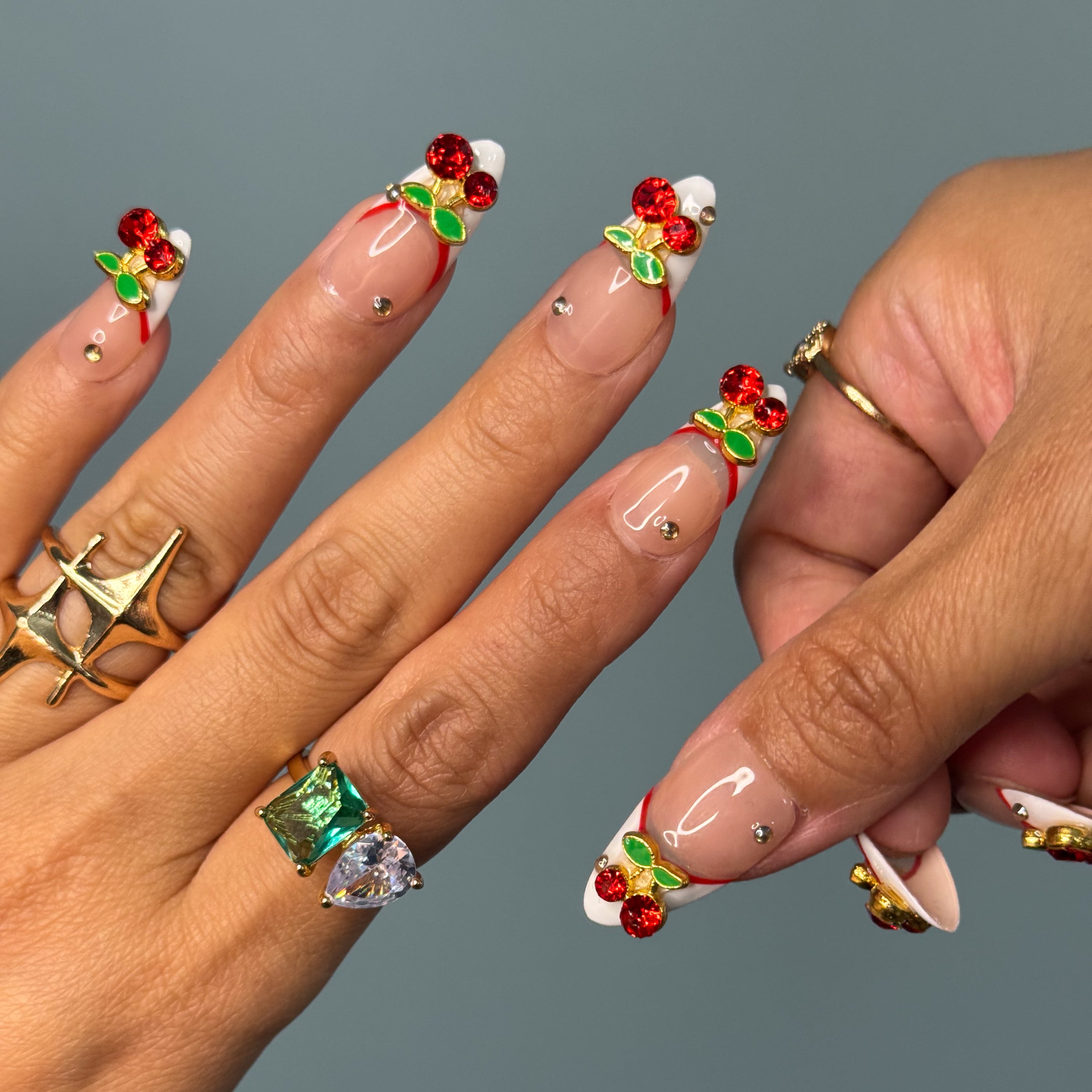 Red French Accent: Classic meets modern with a deep red curve layered over crisp white.
• 3D Cherry Charms: Crystal-studded cherries and metallic green leaves bring a touch of fruity glam.
• Nude Gloss Base: A soft, neutral backdrop keeps the design fresh and wearable.
• Gold Stud Details: Subtle metallic accents add a hint of luxe sparkle.