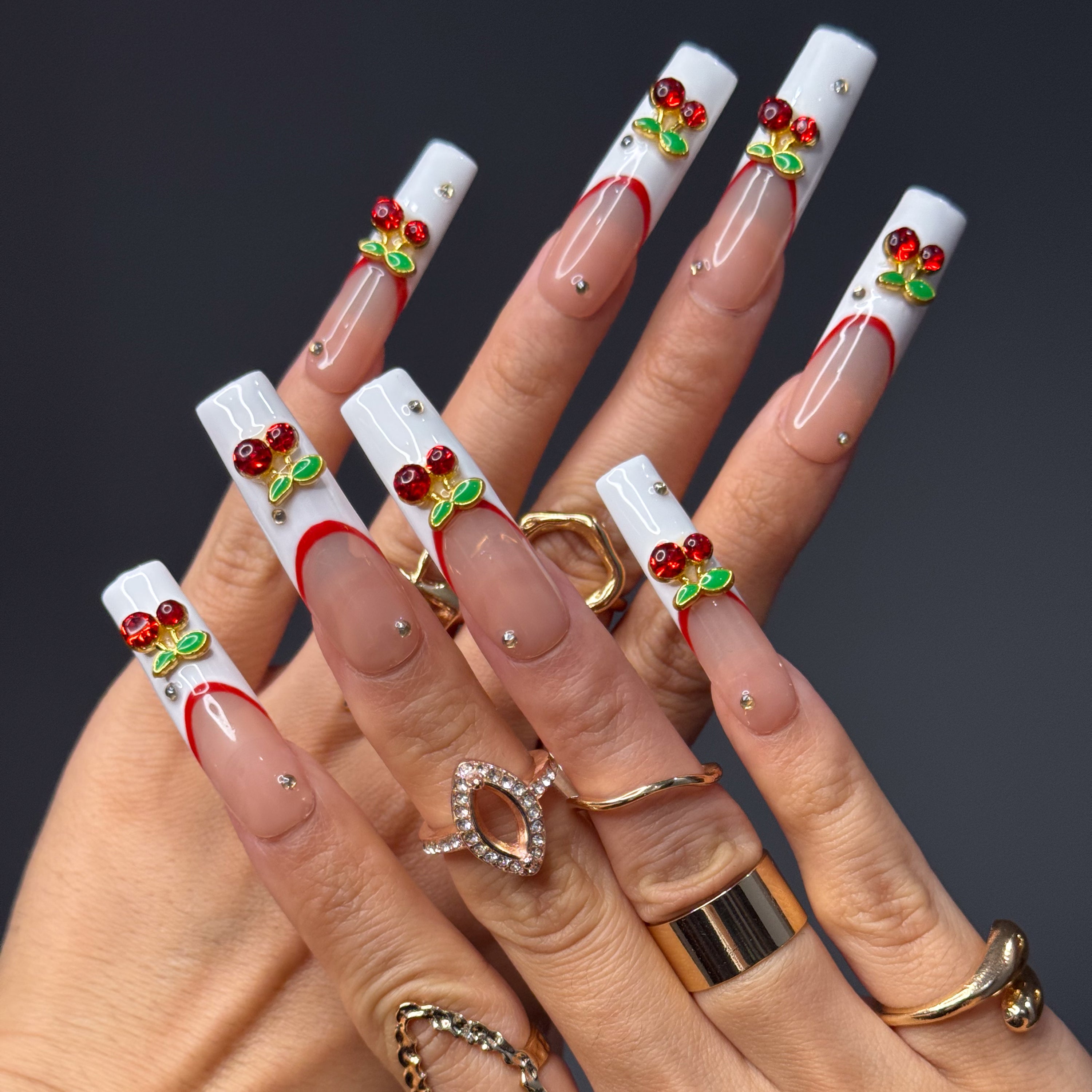 Red French Accent: Classic meets modern with a deep red curve layered over crisp white.
• 3D Cherry Charms: Crystal-studded cherries and metallic green leaves bring a touch of fruity glam.
• Nude Gloss Base: A soft, neutral backdrop keeps the design fresh and wearable.
• Gold Stud Details: Subtle metallic accents add a hint of luxe sparkle.