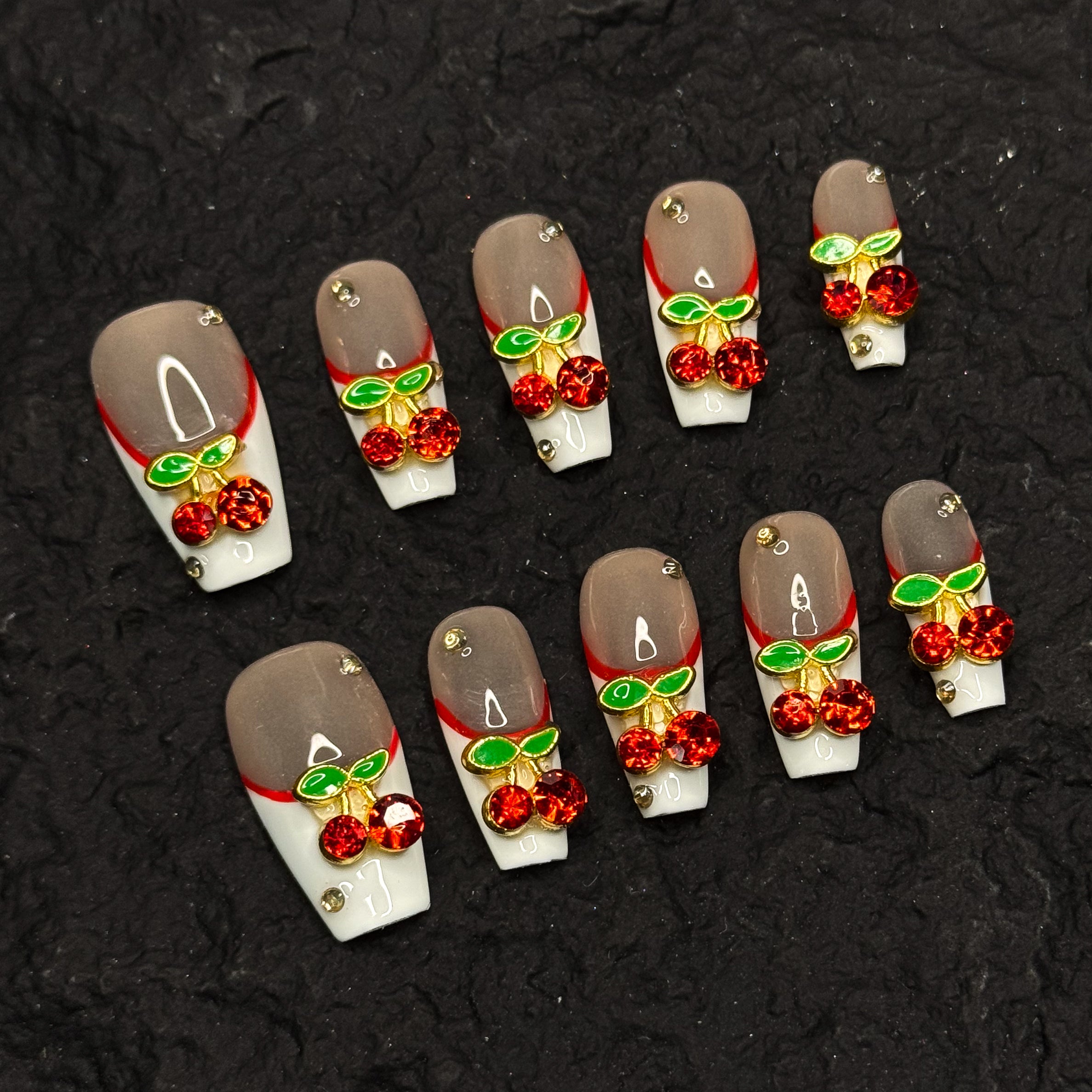 Red French Accent: Classic meets modern with a deep red curve layered over crisp white.
• 3D Cherry Charms: Crystal-studded cherries and metallic green leaves bring a touch of fruity glam.
• Nude Gloss Base: A soft, neutral backdrop keeps the design fresh and wearable.
• Gold Stud Details: Subtle metallic accents add a hint of luxe sparkle.