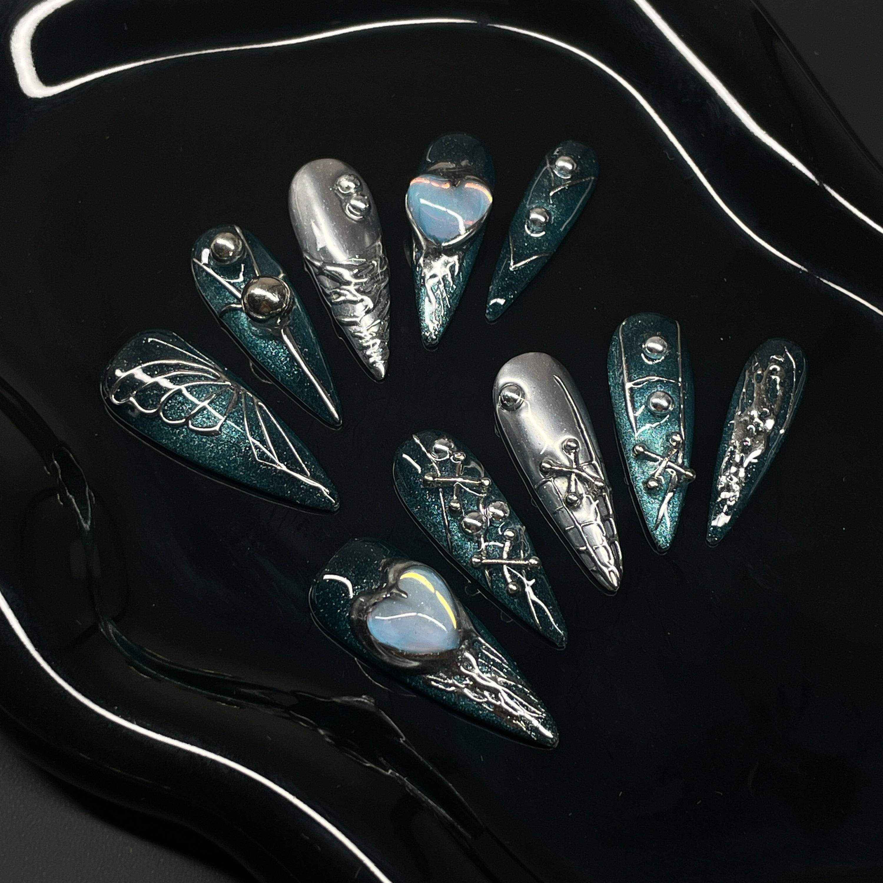 Deep Teal Shimmer: A rich, high-shine base that shifts like oceanic starlight.
• Sculpted Chrome Details: Metallic webs, laces, and barbed textures add mechanical seduction.
• Opal Heart Charms: Centerpieces that glow like tech-enhanced relics of love.
• Mixed Metallic Motifs: Piercing spikes, X details, and dual-tone composition make each nail a statement.
• Long Stiletto Shape: Extra-long, sharply tapered tips designed for digital divas and aesthetic assassins.