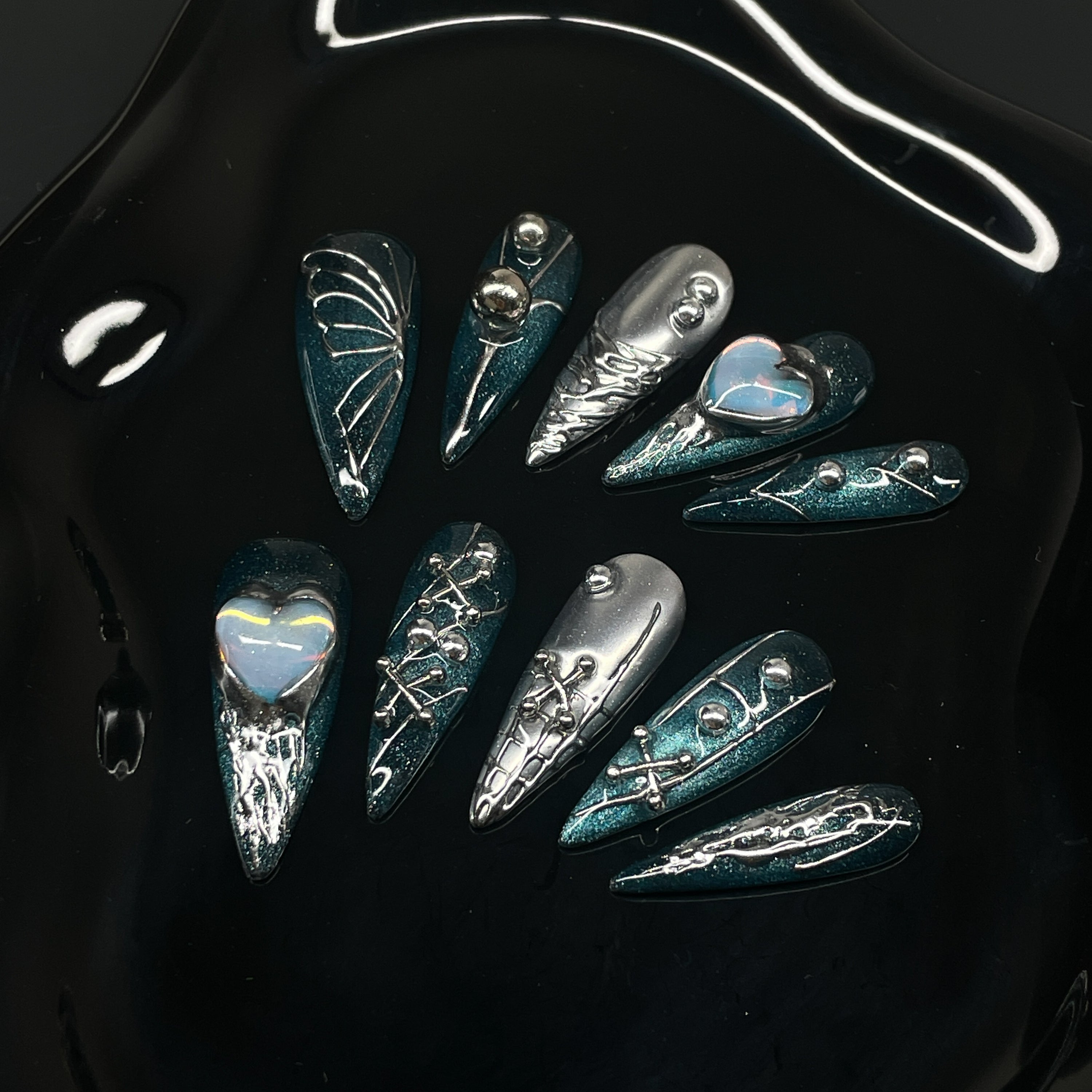 Deep Teal Shimmer: A rich, high-shine base that shifts like oceanic starlight.
• Sculpted Chrome Details: Metallic webs, laces, and barbed textures add mechanical seduction.
• Opal Heart Charms: Centerpieces that glow like tech-enhanced relics of love.
• Mixed Metallic Motifs: Piercing spikes, X details, and dual-tone composition make each nail a statement.
• Long Stiletto Shape: Extra-long, sharply tapered tips designed for digital divas and aesthetic assassins.