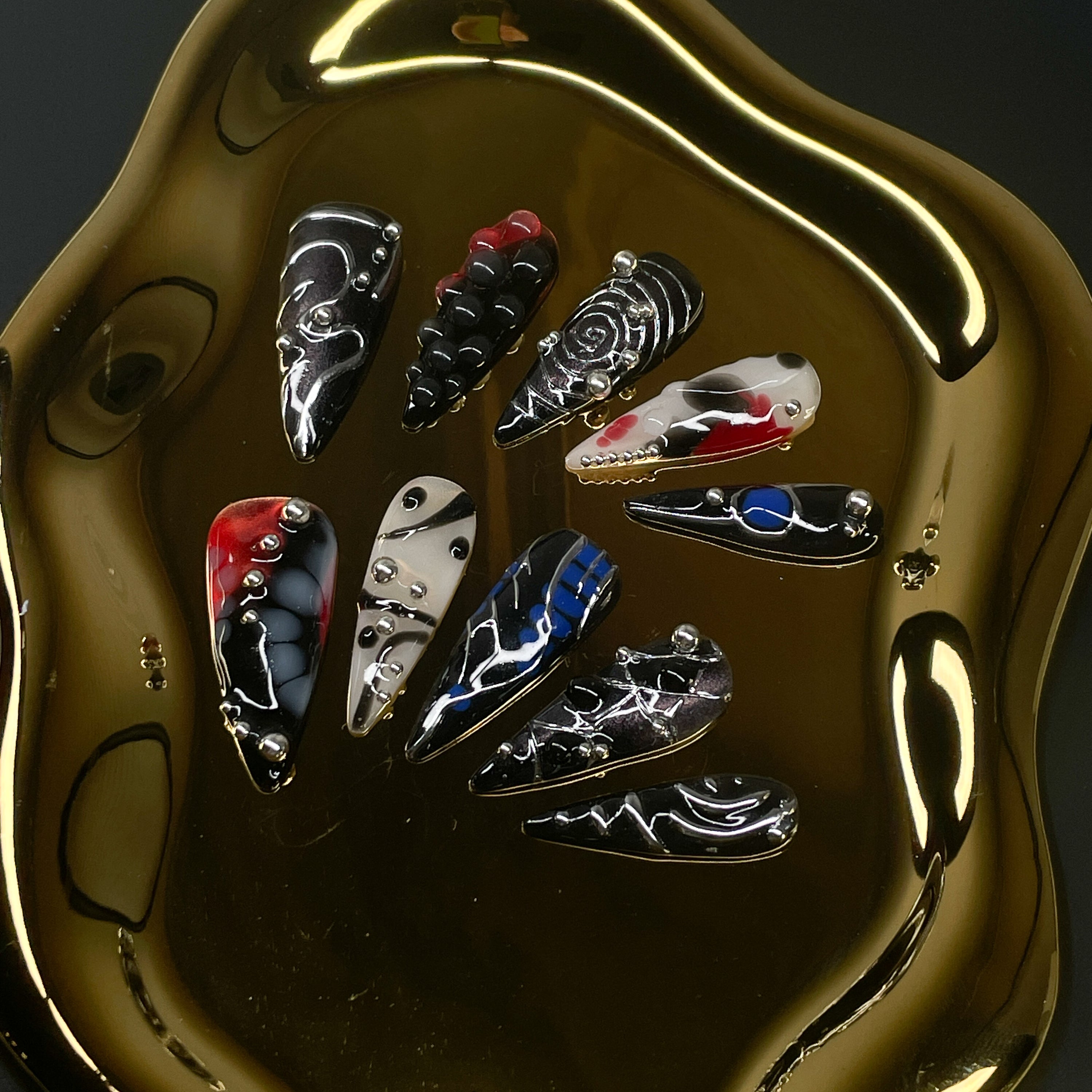 Futuristic Color Clash: Electric blue, blood red, and bone white swirl against chrome black for an ultra-modern contrast. 3D Sculptural Texture: Raised silver spheres and layered gel details create depth and motion. Glitch Art Motifs: Bold linework, chaotic patterns, and fragmented symmetry evoke digital distortion. Metallic Mirror Finish: Sleek chrome effects reflect light like machine parts in motion. Stiletto Shape: Fiercely pointed tips enhance the dramatic, cybernetic aesthetic—made to cut through both
