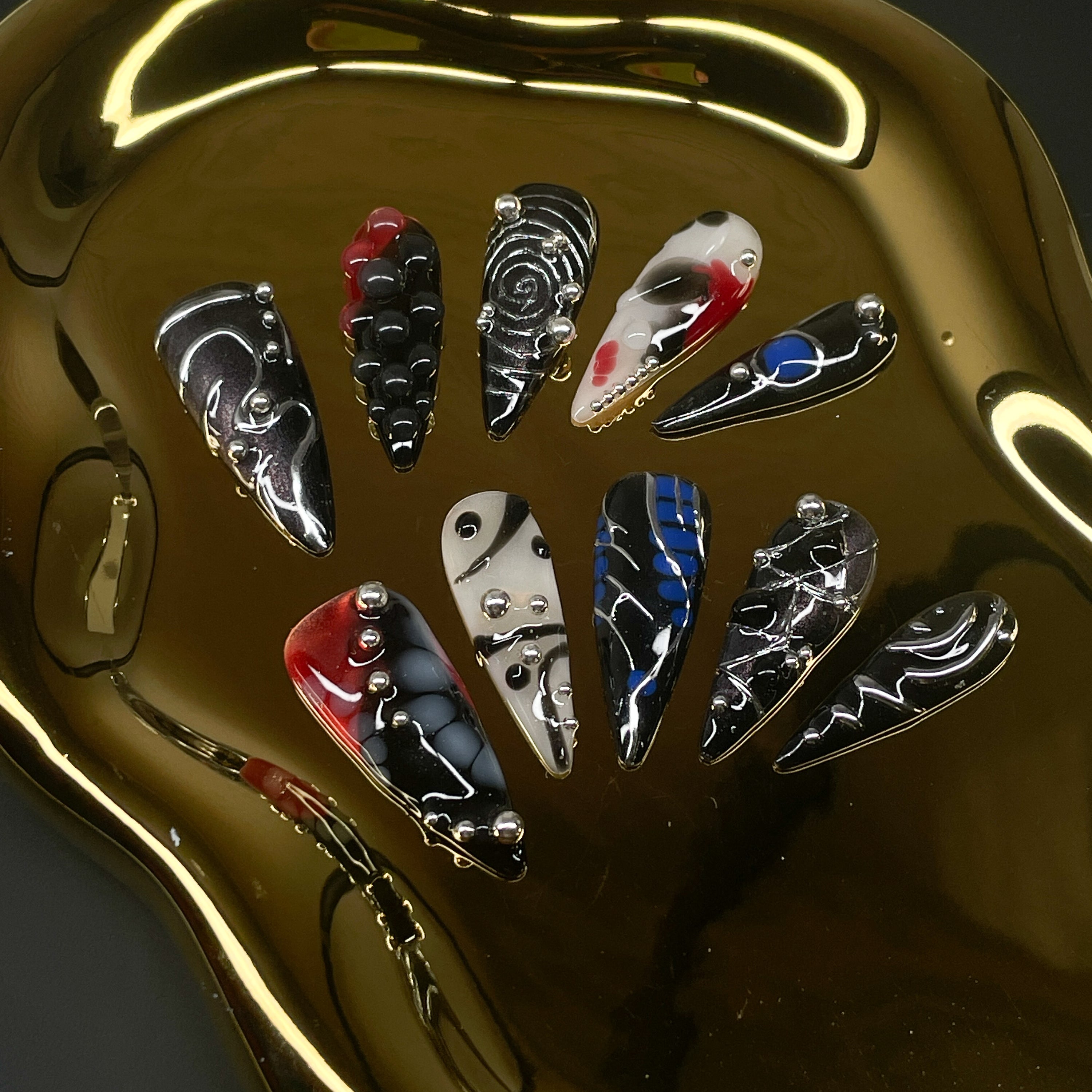 Futuristic Color Clash: Electric blue, blood red, and bone white swirl against chrome black for an ultra-modern contrast. 3D Sculptural Texture: Raised silver spheres and layered gel details create depth and motion. Glitch Art Motifs: Bold linework, chaotic patterns, and fragmented symmetry evoke digital distortion. Metallic Mirror Finish: Sleek chrome effects reflect light like machine parts in motion. Stiletto Shape: Fiercely pointed tips enhance the dramatic, cybernetic aesthetic—made to cut through both