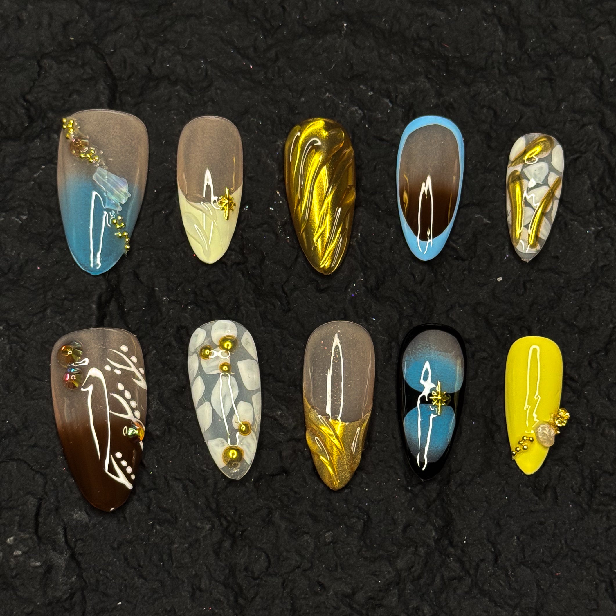 Warm Earth Palette: Deep browns, sandy taupes, mustard yellows, and rich sky blues for a grounded, boho-luxe vibe. Metallic Gold Accents: Molten chrome details and sculpted lines mimic mineral veins and glowing rock strata. Abstract Nature Patterns: Painted pebble motifs, fluid swirls, and organic forms evoke raw, artisanal textures. Beaded Embellishments: Tiny gold beads and crystal touches add luxe detail without overpowering. Almond Shape: An elegant, tapered silhouette that balances artistic drama with 