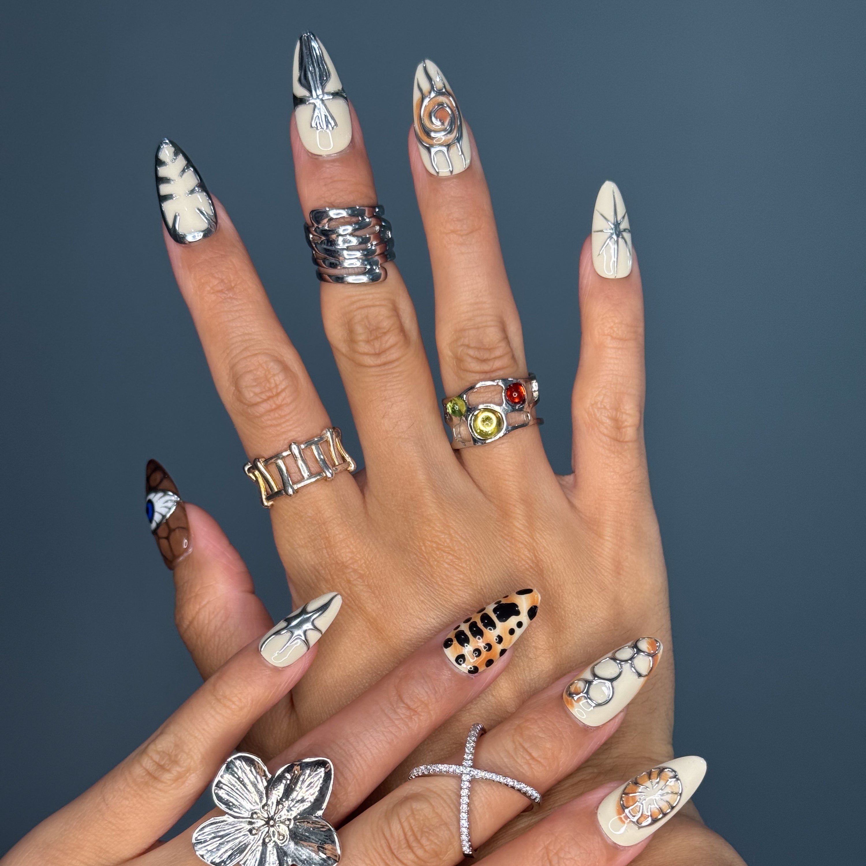 Earthy Neutrals: A palette of caramel, bone, clay, and soft browns grounded in organic beauty.
• Ancient Symbols: Spirals, stars, shells, and skeletal imprints rendered in molten silver for a modern twist.
• Textured 3D Elements: Sculpted chrome and layered gel create tactile depth that mimics stone carvings.
• Tribal Sea Motifs: From fish scales to squid silhouettes and protective eyes, each nail tells a primordial story.
• Almond Shape: A smooth, natural curve that complements the fossil-inspired designs 