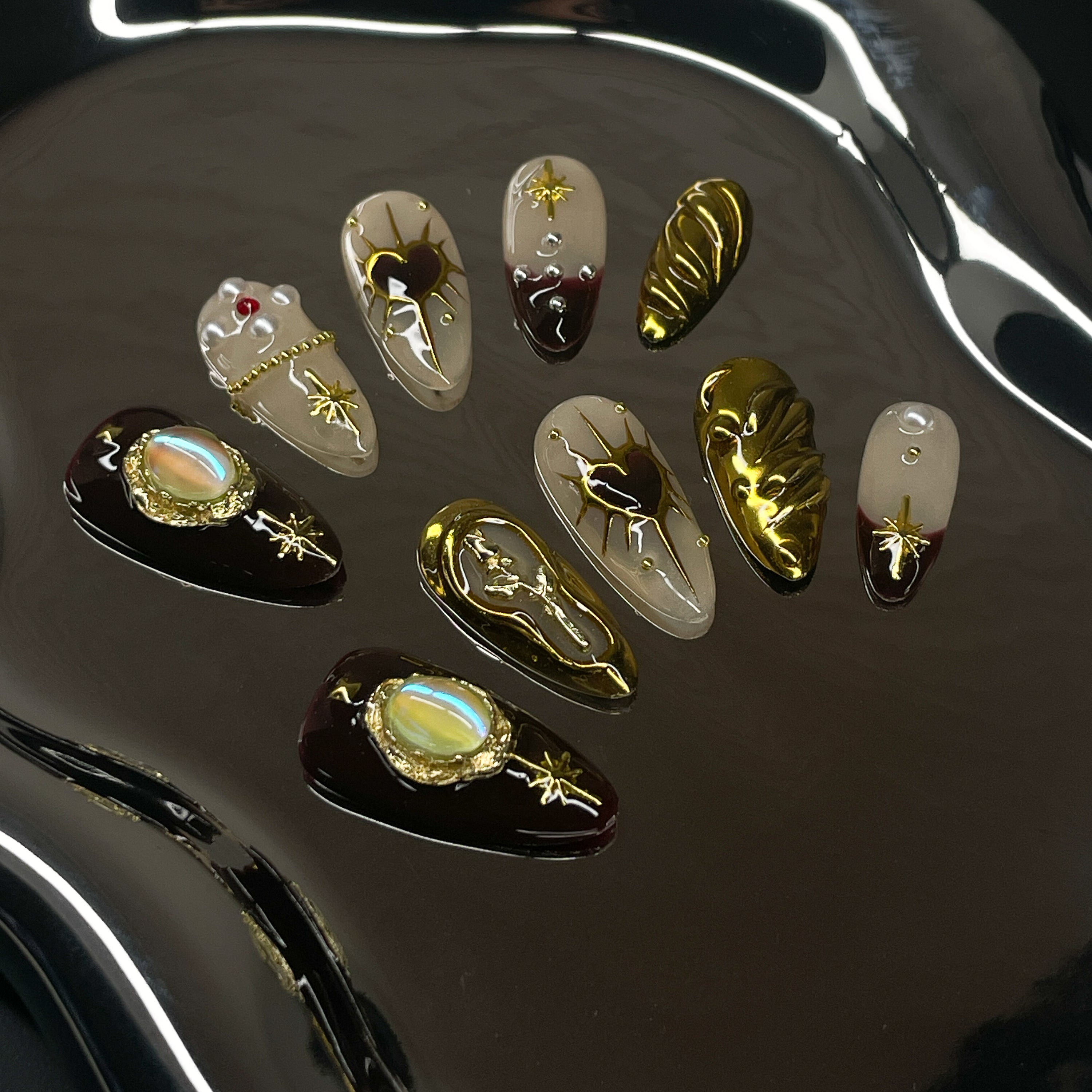  Regal Color Palette: Nude and burgundy tones kissed with rich gold and soft shimmer.
• 3D Sacred Elements: Raised heart motifs, golden auras, and vintage-inspired opal charms for heavenly detail.
• Luxe Metallic Accents: Sculpted gold swirls, chain links, and stars add depth and decadence.
• Mixed Textures: Smooth nudes contrast with glossy dark tips and radiant metallic overlays.
• Almond Shape: A soft, elegant silhouette that enhances the baroque, relic-like beauty.