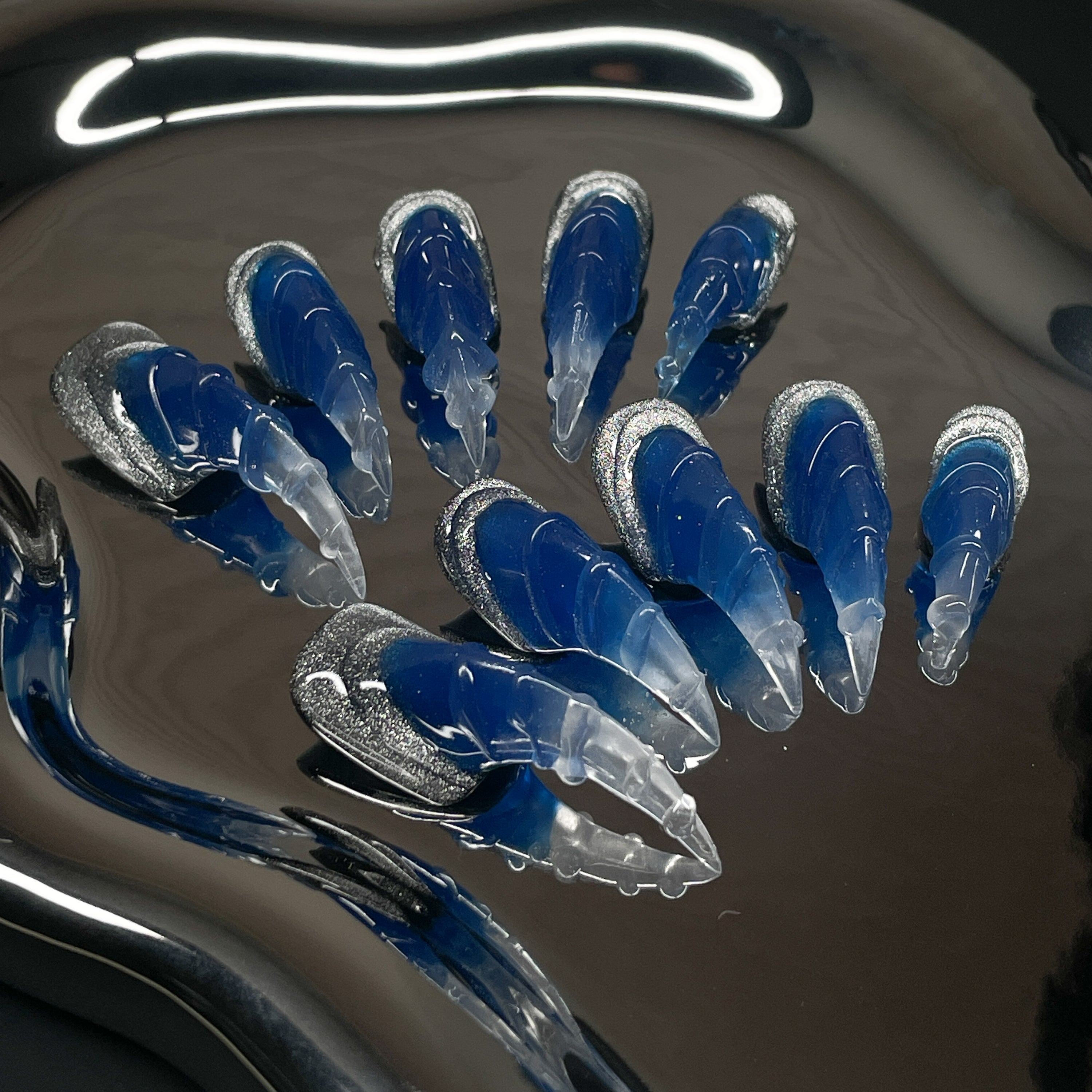Glacier Blue Ombre: Frosty blues fade into translucent crystal tips like ice breaking over deep water.
• 3D Claw Sculpt: Each nail is shaped with fierce, curved ridges for a wild, talon-like silhouette.
• Silver Ice Accents: Glittery chrome edges mimic frozen crusts and shimmering snow.
• Frosted Finish: A mix of matte and gloss textures gives this set the ultimate arctic edge.
• Claw Shape: Sharp, sculpted tips that extend like icy weapons—bold, edgy, and unapologetic.
