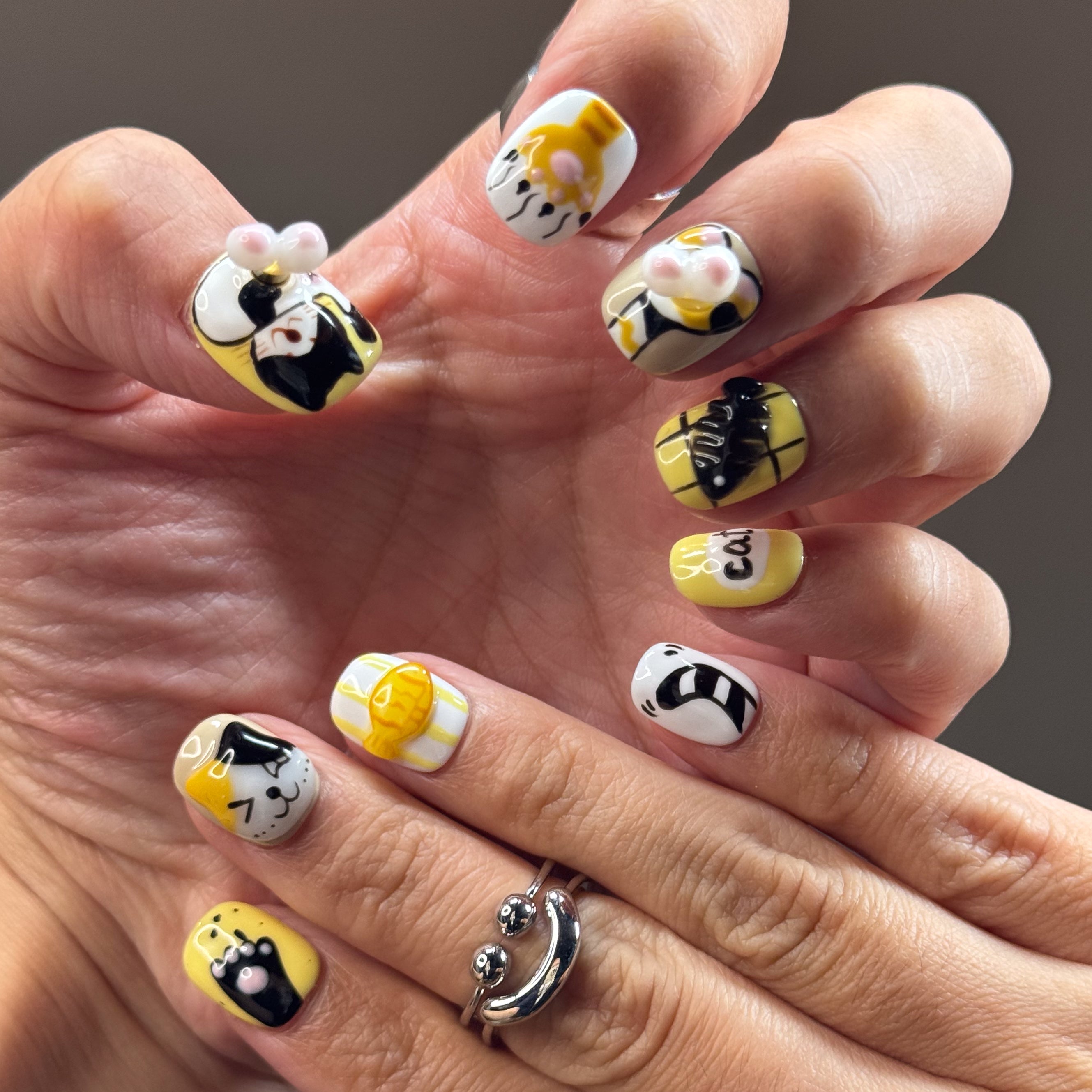 • Adorable Cat Art: Hand-painted kitty faces in orange and calico with the cutest expressions.
• 3D Paw Details: Puffy jellybean paw pads you’ll want to boop all day.
• Playful Motifs: Fish bones, yarn balls, and the word “cat” in bold black for extra whimsy.
• Cheerful Colors: A happy palette of yellow, orange, white, and black.
• Short Square Shape: Easy to wear daily while keeping the design bold and fun.