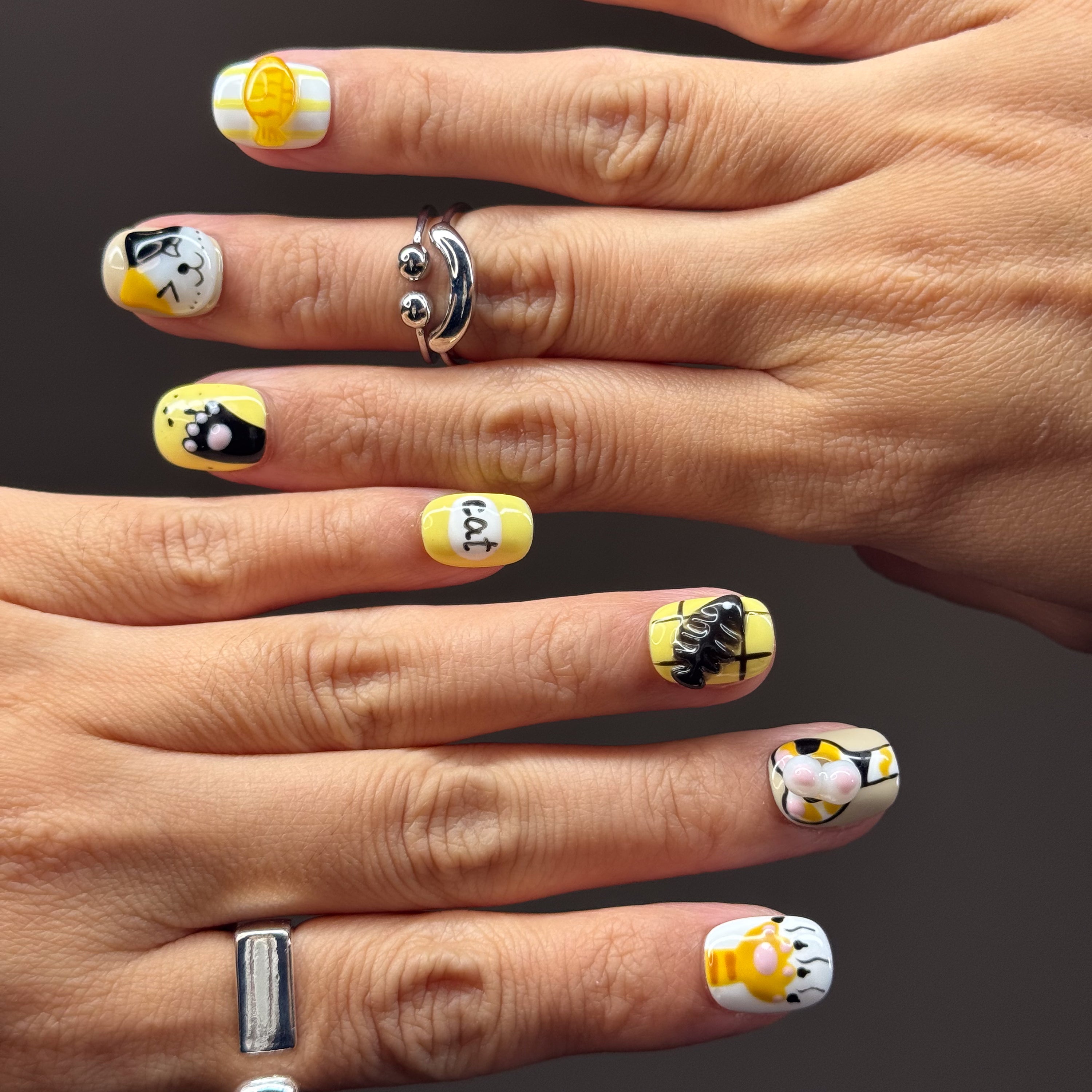 • Adorable Cat Art: Hand-painted kitty faces in orange and calico with the cutest expressions.
• 3D Paw Details: Puffy jellybean paw pads you’ll want to boop all day.
• Playful Motifs: Fish bones, yarn balls, and the word “cat” in bold black for extra whimsy.
• Cheerful Colors: A happy palette of yellow, orange, white, and black.
• Short Square Shape: Easy to wear daily while keeping the design bold and fun.