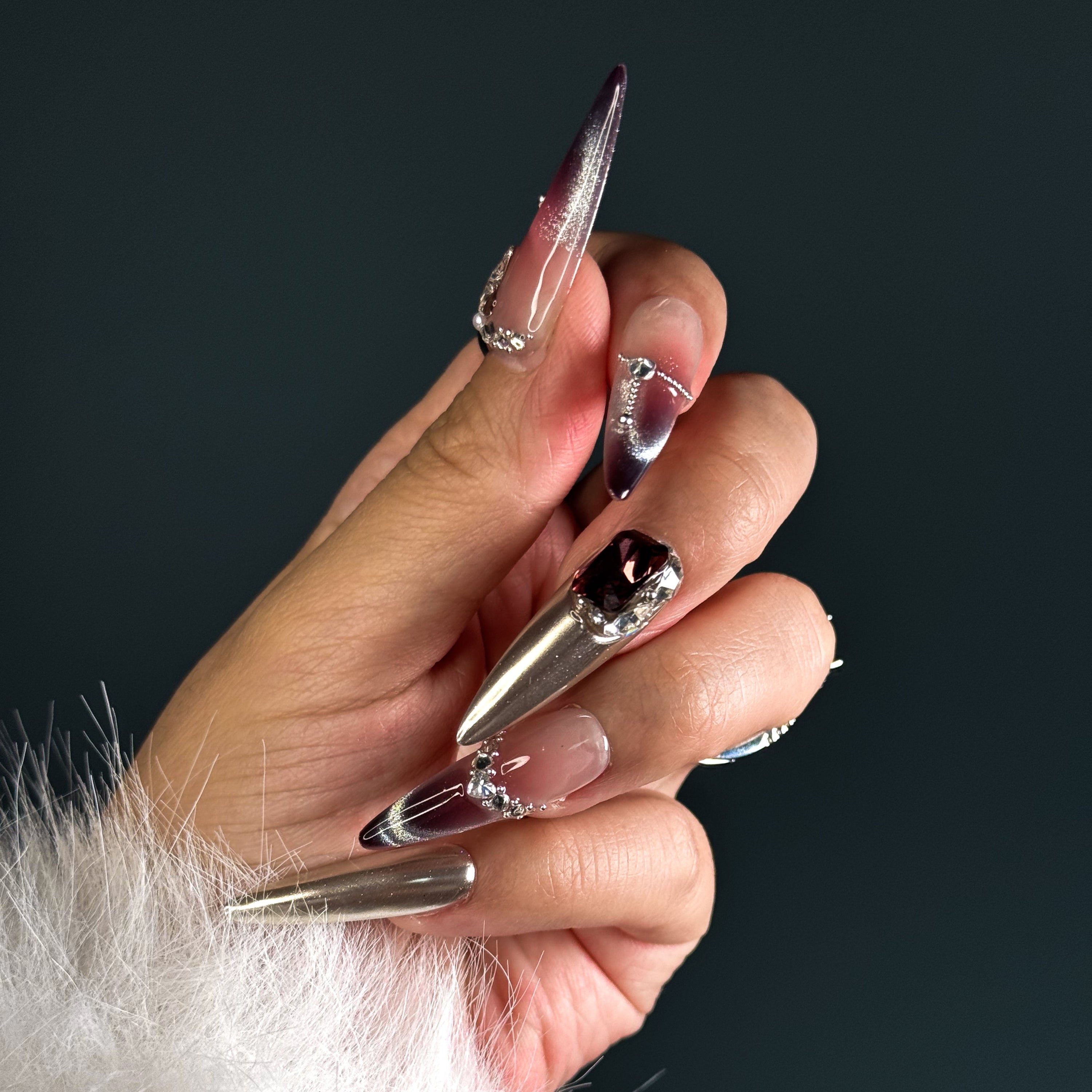 Indulge in pure luxury with Royal Mirage Press-On Nails. These extra-long stiletto nails feature a breathtaking purple ombre blended with silver, creating a regal and enchanting look. Accented with dazzling diamonds and gemstone decorations, each nail sparkles with elegance. A mirror-like full silver nail adds a bold, high-fashion edge, while the cat-eye effect enhances the depth and mystery of the design.