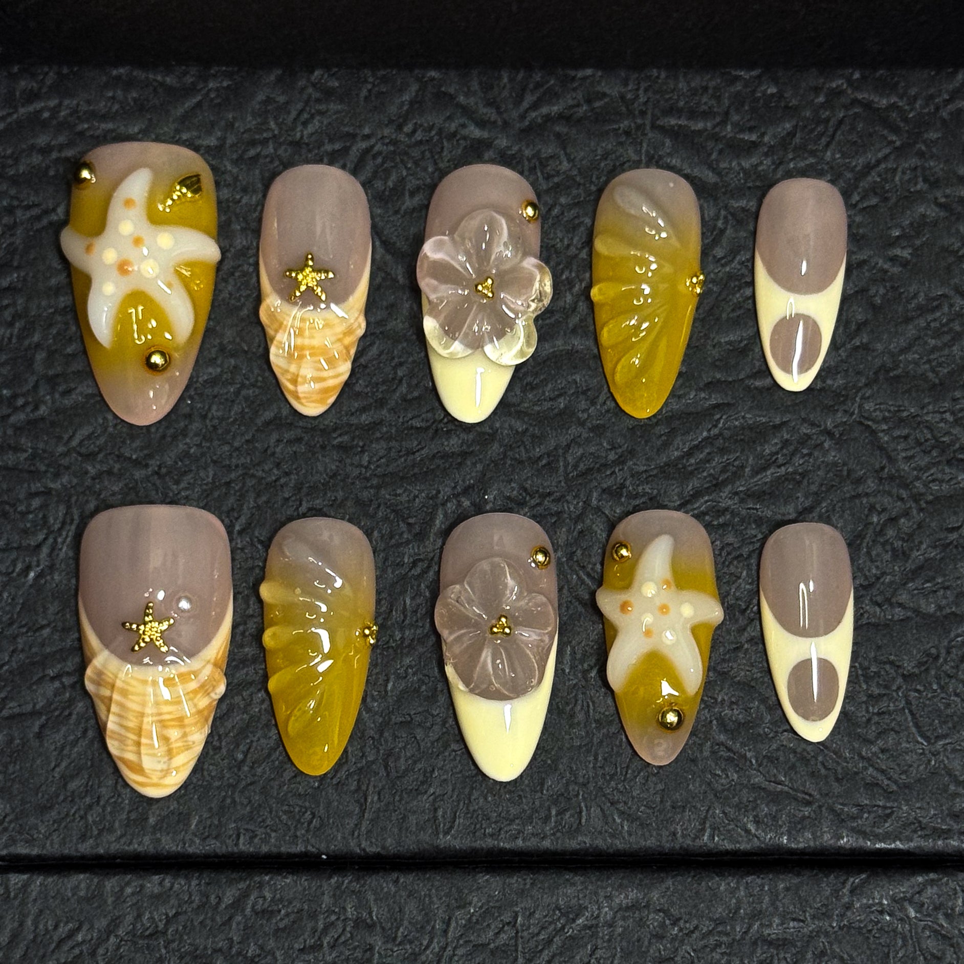Coastal Color Palette: Taupe, honey yellow, ivory cream, and shell-inspired swirls for that sun-bleached vibe.
• 3D Starfish & Flowers: Hand-sculpted ocean motifs bring depth and summer romance to each nail.
• Textured Shell Accents: Wave-like and seashell textures evoke the feeling of walking barefoot on warm sand.
• Gold Bead Embellishments: Delicate studs and tiny stars add a glimmer of ocean treasure.
• Almond Shape: Natural, beachy elegance with a soft tapered point perfect for sunny adventures.