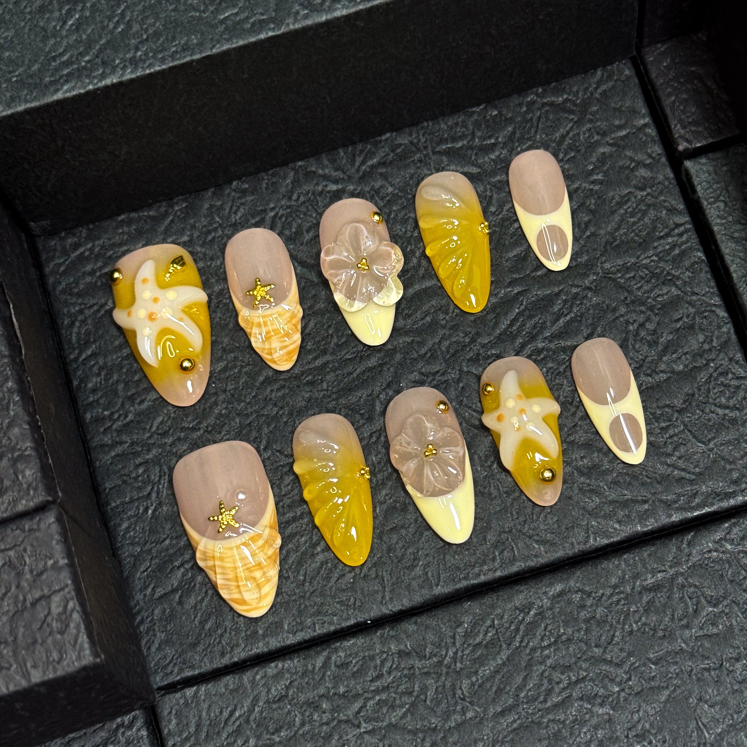 Coastal Color Palette: Taupe, honey yellow, ivory cream, and shell-inspired swirls for that sun-bleached vibe.
• 3D Starfish & Flowers: Hand-sculpted ocean motifs bring depth and summer romance to each nail.
• Textured Shell Accents: Wave-like and seashell textures evoke the feeling of walking barefoot on warm sand.
• Gold Bead Embellishments: Delicate studs and tiny stars add a glimmer of ocean treasure.
• Almond Shape: Natural, beachy elegance with a soft tapered point perfect for sunny adventures.