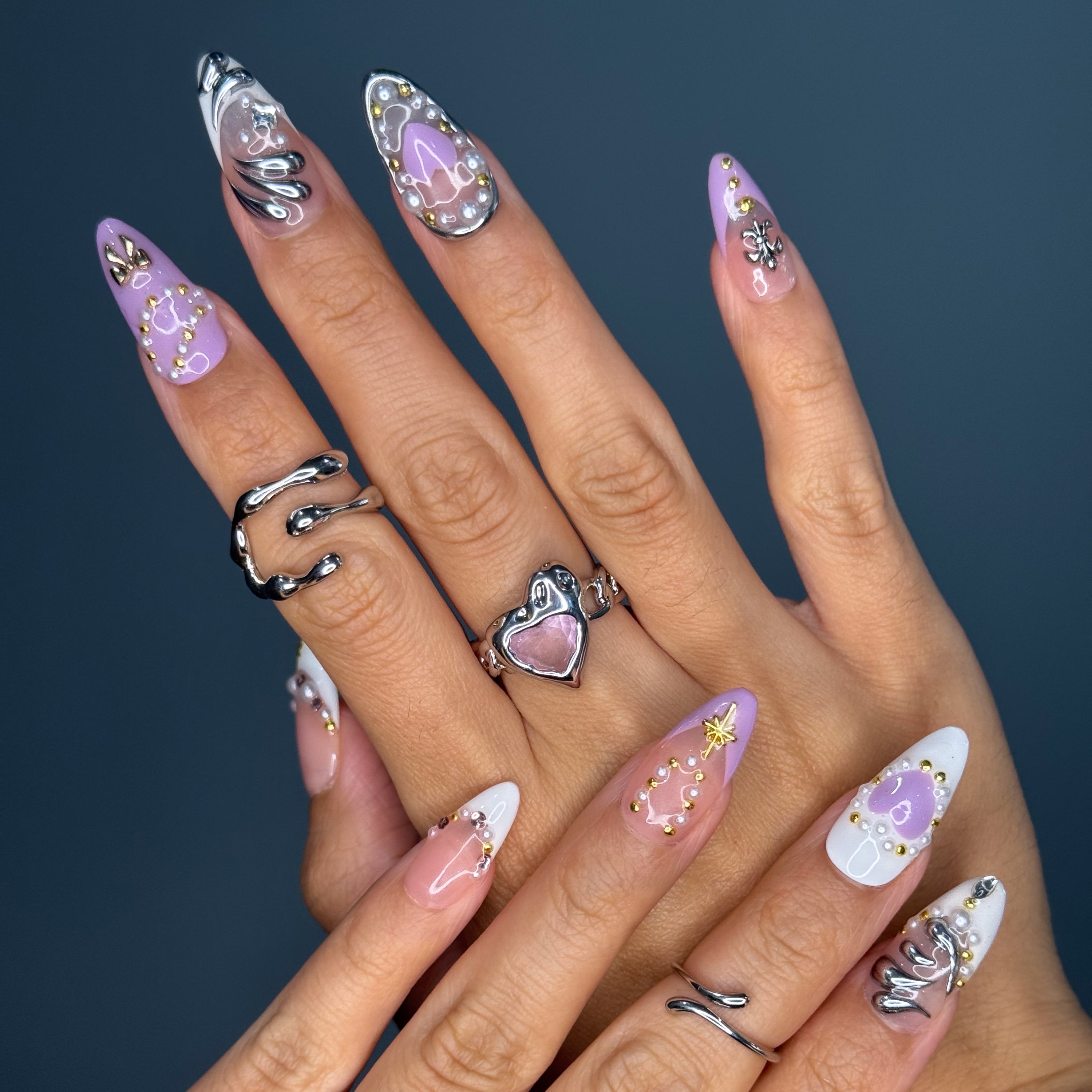 Soft Romantic Tones: A dreamy palette of dusty pink, cocoa brown, ivory, and lavender.
• 3D Heart & Chain Details: Sculpted hearts, pearls, and golden charms give a love-me-but-don’t-cross-me energy.
• Chrome Highlights: Shiny silver and gold accents add edge to the softness.
• Mixed Motifs: Features butterflies, barbed wire, and celestial symbols—where dreamy meets dangerous.
• Almond Shape: Flattering and elegant, this shape balances delicate charm with fierce style.