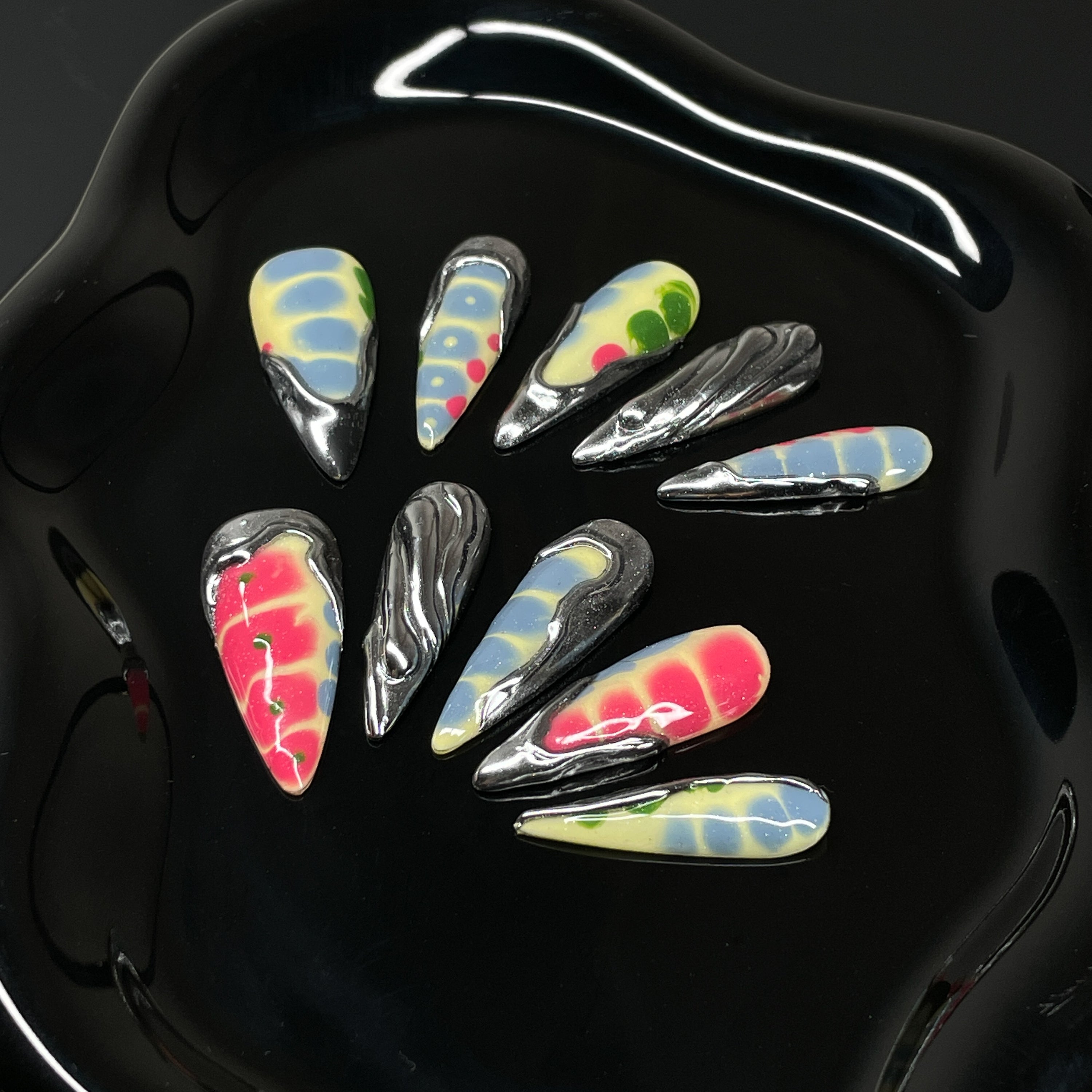Acid Tie-Dye: Vivid blends of yellow, red, blue, and green with smooth, melty gradients.
• Liquid Chrome Accents: Sculptural silver forms create a venomous drip effect that oozes cool.
• 3D Texture Play: Each nail features raised details and dimensional overlays for maximum impact.
• Psychedelic Patterns: Organically chaotic shapes give each tip its own rebellious attitude.
• Long Stiletto Shape: Sharp, elongating points perfect for a fierce, high-fashion finish.