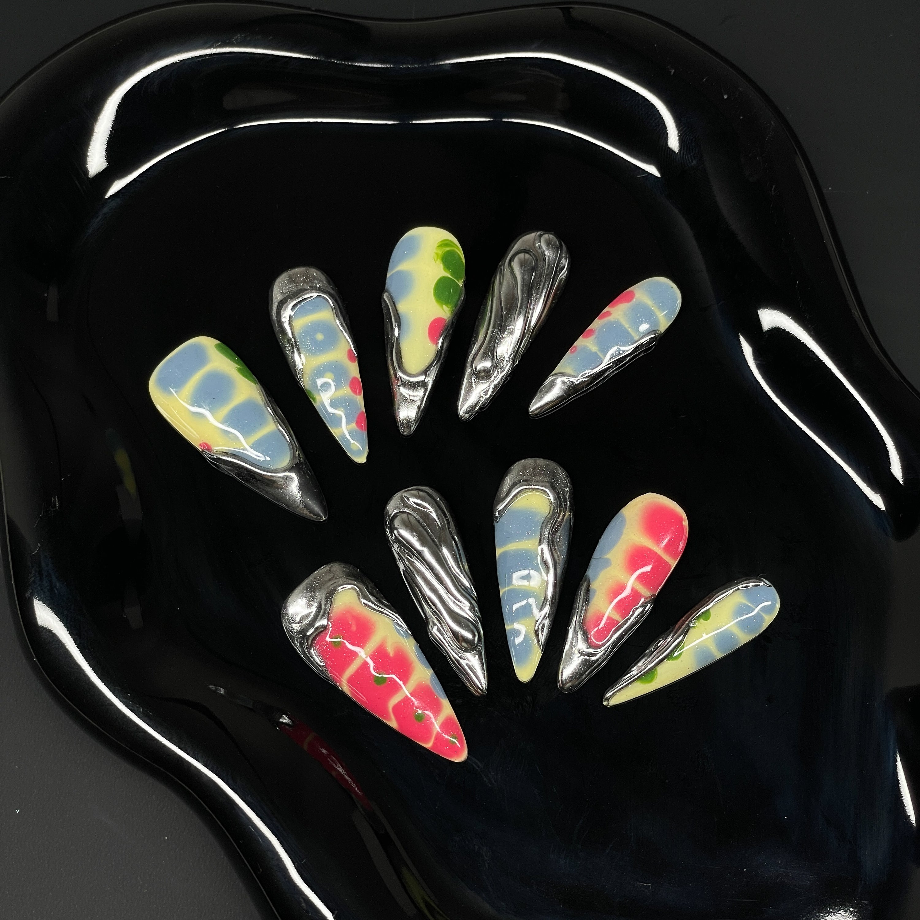 Acid Tie-Dye: Vivid blends of yellow, red, blue, and green with smooth, melty gradients.
• Liquid Chrome Accents: Sculptural silver forms create a venomous drip effect that oozes cool.
• 3D Texture Play: Each nail features raised details and dimensional overlays for maximum impact.
• Psychedelic Patterns: Organically chaotic shapes give each tip its own rebellious attitude.
• Long Stiletto Shape: Sharp, elongating points perfect for a fierce, high-fashion finish.