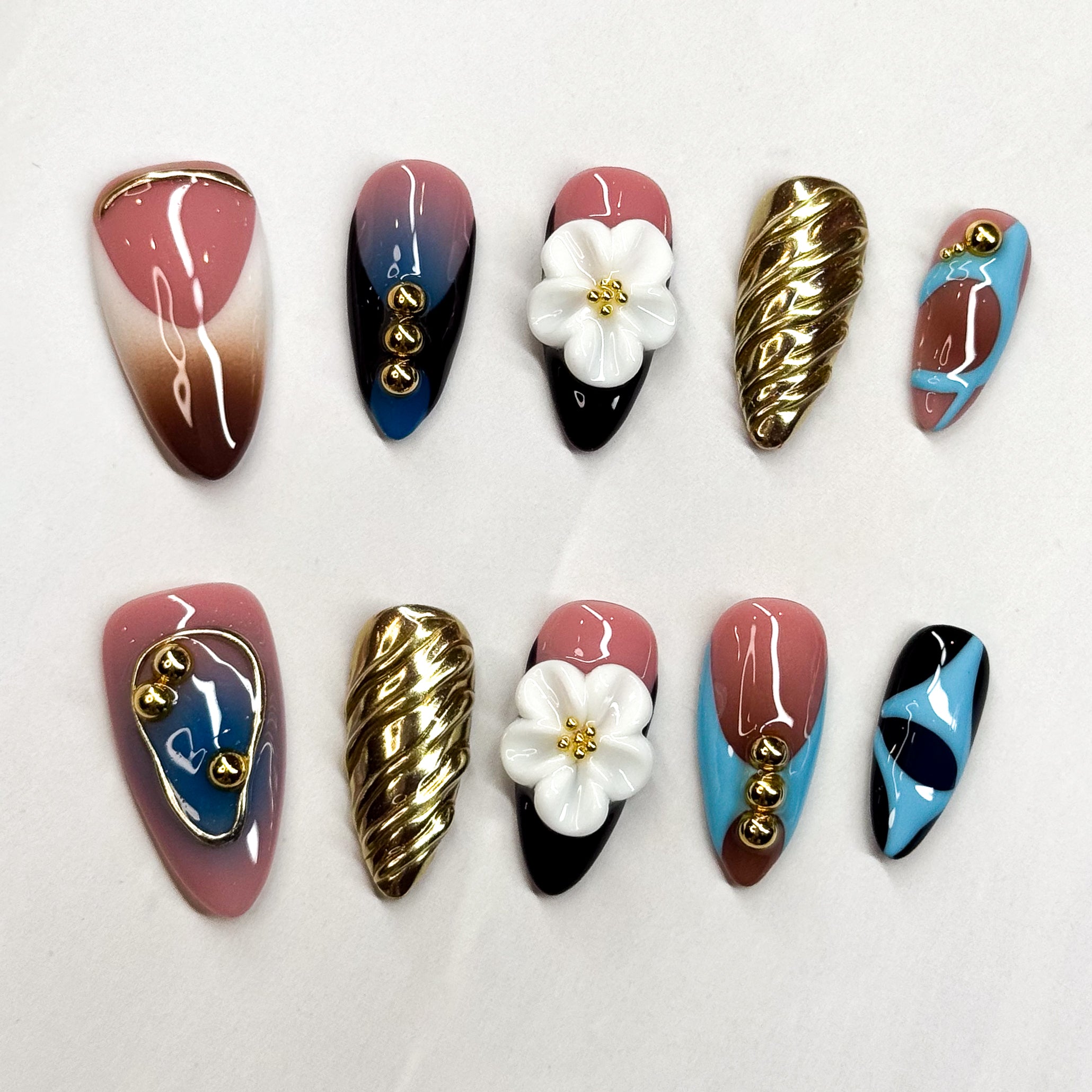 • Bold Design: A blend of pink, blue, black, and brown with gold metallic details.
• Artistic Accents: 3D sculpted flower charms and twisted gold textures.
• Playful Abstracts: Hand-painted patterns and high-shine finishes.
• Unique Pairing: Each nail is designed as a statement, yet together they create a harmonious, modern look.
• Handmade & Reusable: Premium 3D gel craftsmanship ensures durability and reusability.