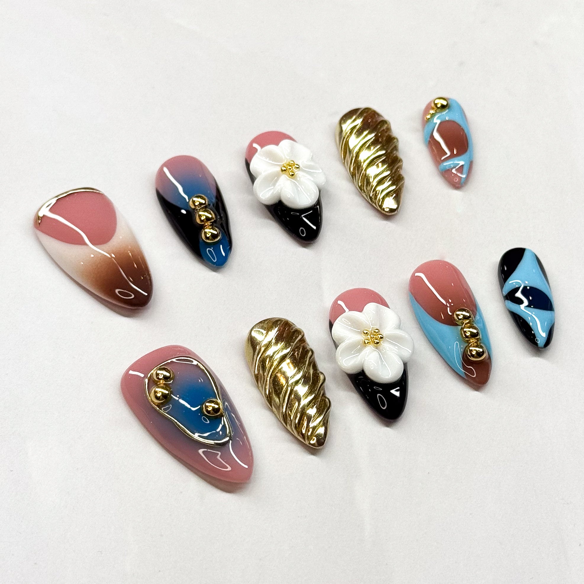 • Bold Design: A blend of pink, blue, black, and brown with gold metallic details.
• Artistic Accents: 3D sculpted flower charms and twisted gold textures.
• Playful Abstracts: Hand-painted patterns and high-shine finishes.
• Unique Pairing: Each nail is designed as a statement, yet together they create a harmonious, modern look.
• Handmade & Reusable: Premium 3D gel craftsmanship ensures durability and reusability.