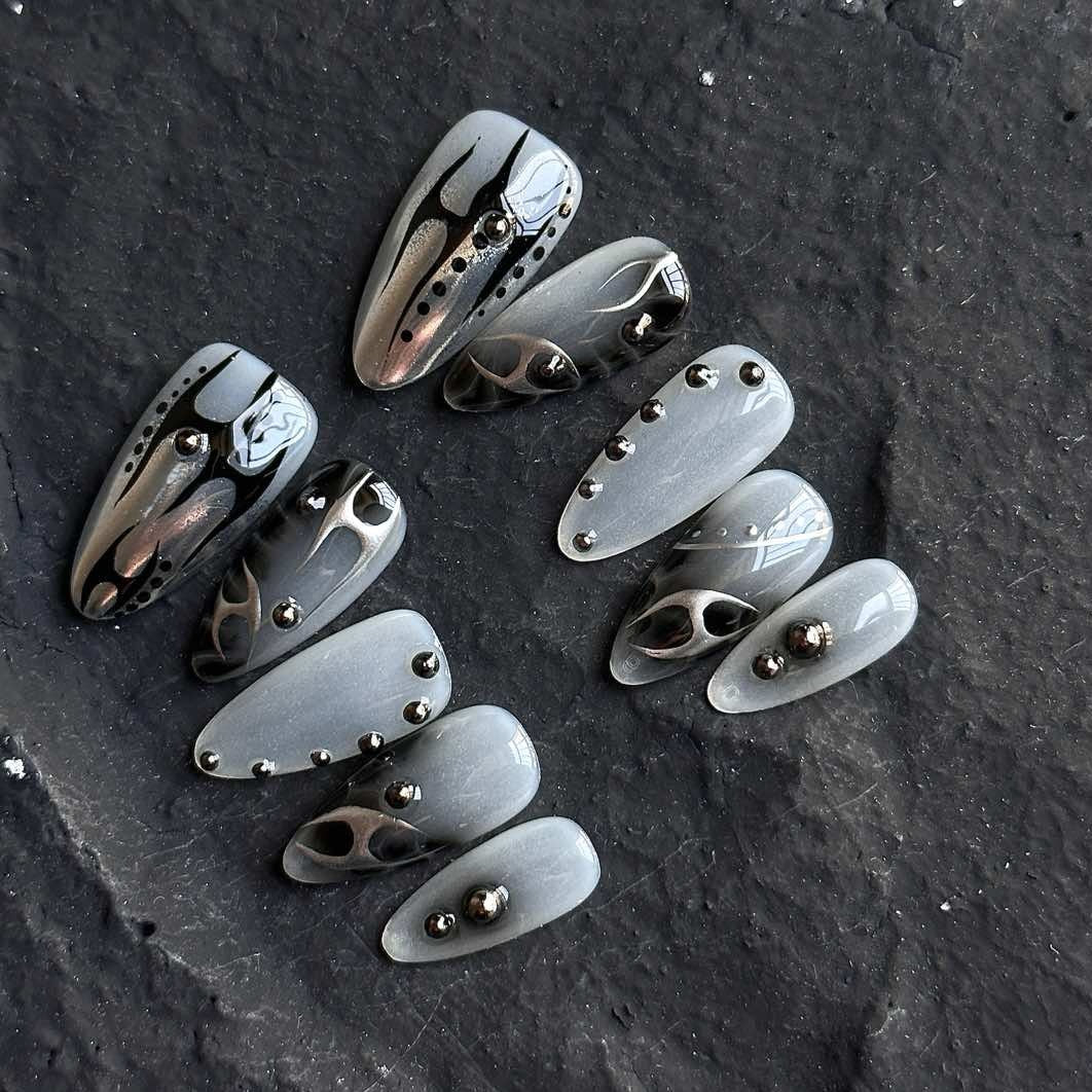 Unleash a bold, futuristic edge with Obsidian Chrome Press-On Nails. This striking set features a smoky gray base with sleek black chrome flame designs, accented by metallic studs for a modern, industrial-inspired aesthetic. The combination of glossy and matte finishes, along with sculpted 3D elements, creates a daring and sophisticated look that’s perfect for trendsetters who love an avant-garde touch.