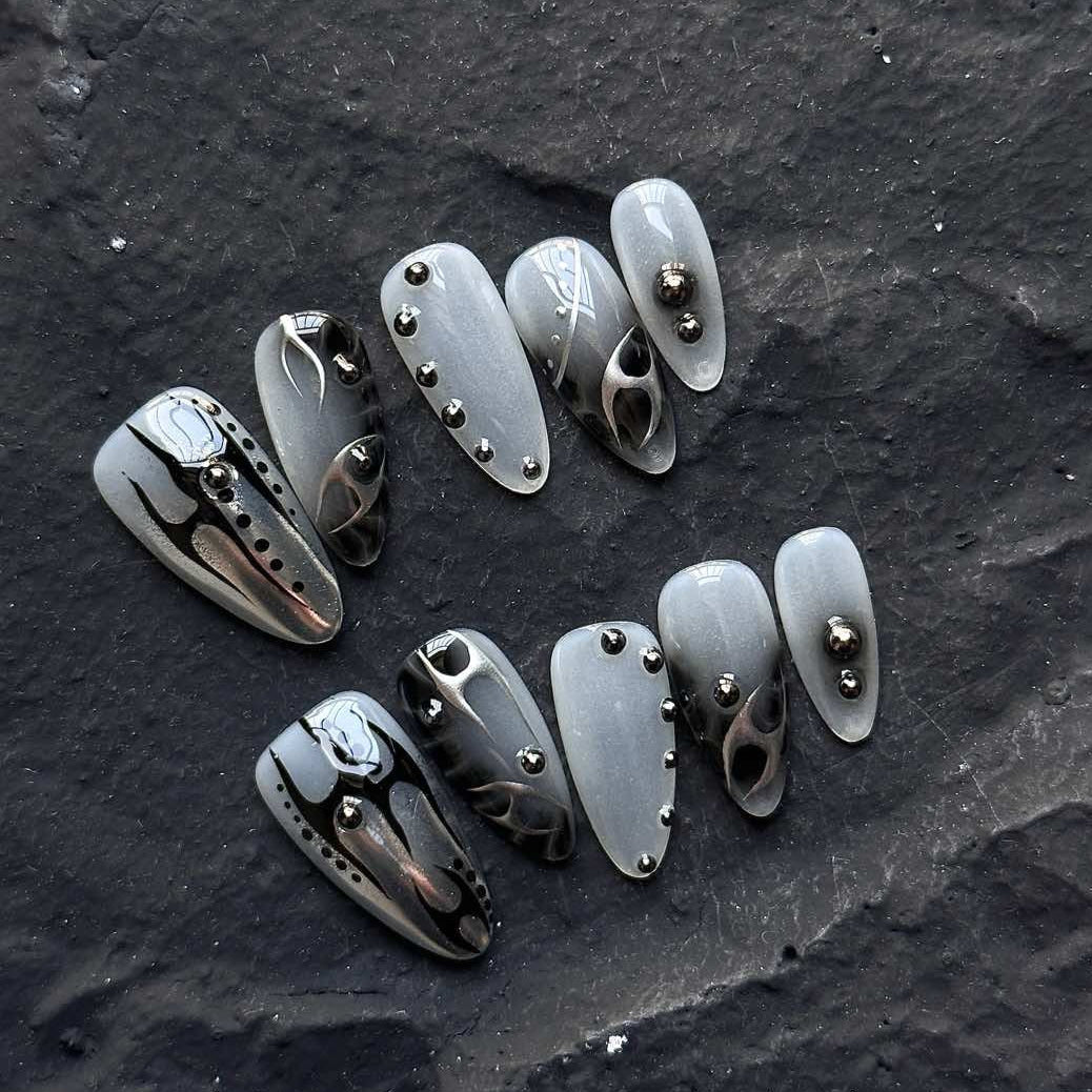 Unleash a bold, futuristic edge with Obsidian Chrome Press-On Nails. This striking set features a smoky gray base with sleek black chrome flame designs, accented by metallic studs for a modern, industrial-inspired aesthetic. The combination of glossy and matte finishes, along with sculpted 3D elements, creates a daring and sophisticated look that’s perfect for trendsetters who love an avant-garde touch.